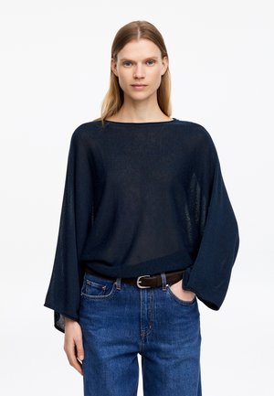 Žena s rovnými blond vlasmi nosí voľný tmavomodrý sveter so širokými rukávmi a modré džínsy, stojí s jednou rukou v kapse.