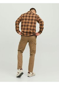 Camicia a quadri marrone e beige con polsini a bottone indossata sopra una maglietta marrone, abbinata a pantaloni cargo beige e sneakers bianche con accenti rossi.