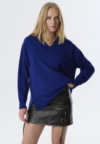 Blauer V-Ausschnitt-Pullover aus weichem Material mit langen Ärmeln, kombiniert mit einem schwarzen Minirock aus Kunstleder, der über Reißverschlüsse und Taschen verfügt.