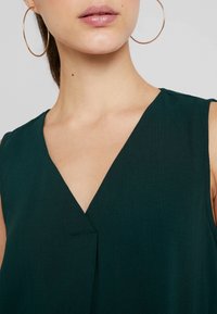 Dunkelgrünes ärmelloses Oberteil mit V-Ausschnitt, das eine subtile Textur und eine zentrale Faltenlage aufweist. Große goldene Creolen sind sichtbar.