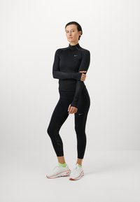Nike sort langærmet bluse med lynlås-krave, der har teksturerede detaljer på ærmerne, parret med sorte leggings og hvide træningssko.