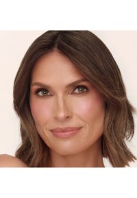 Primo piano di un volto con capelli castano medio e mossi, occhi verdi e trucco leggero caratterizzato da un delicato fard rosa e un colore di labbra nude, su uno sfondo neutro.