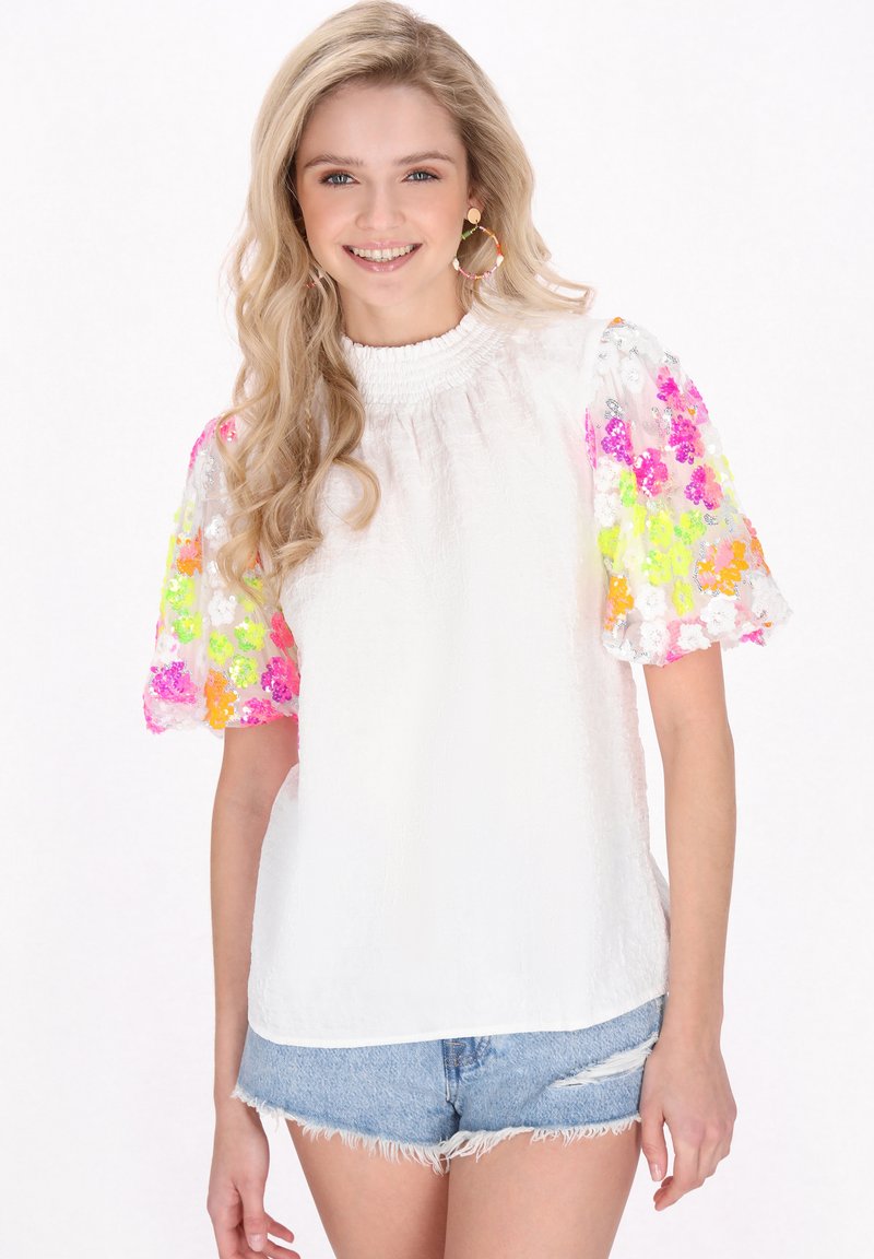 Hvid top med smocket halsudskæring og korte, gennemsigtige ærmer pyntet med pink, orange og grønne blomster-sekvenser. Matchet med denimshorts.