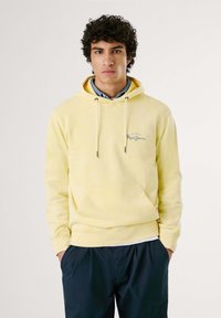 Felpa gialla con tasca frontale e cappuccio regolabile, presenta un logo blu sul petto, indossata sopra una camicia colletto azzurro.