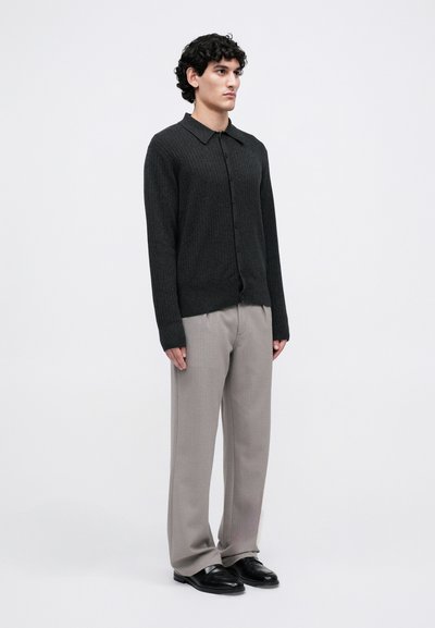 Uomo che indossa un cardigan a coste scuro con bottoni, pantaloni larghi grigio chiaro e scarpe nere, in piedi contro uno sfondo bianco semplice.