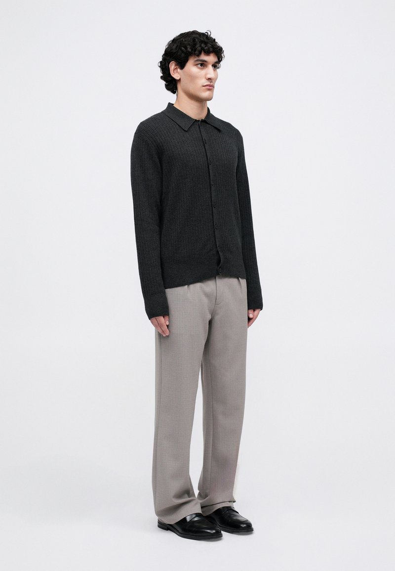 Man wearing dark ribbed button-up cardigan, light gray loose trousers, and black shoes, standing against a plain white background.