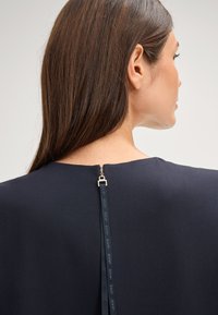 Marineblauwe jurk gezien vanaf de achterkant, met een verticale ritssluiting met een gouden trekker en een merkribbel detail. Glad materiaal zonder zichtbare patronen.