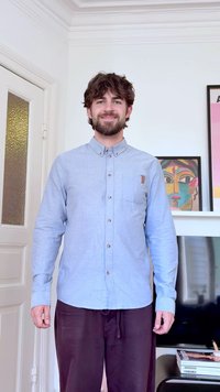 Muškarac s tamnom kosom i bradom, odjeven u svijetloplavu košulju s gumbima i tamne hlače, stoji u svijetloj sobi s uokvirenim umjetničkim djelima i televizorom.