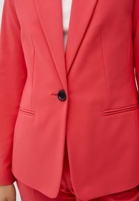 Korálový blazer s jedním černým knoflíkem, klopami a bočními kapsami. Vyrobeno z hladkého materiálu s přiléhavým střihem.