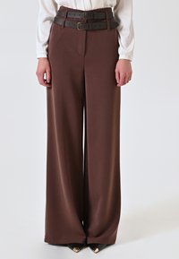 Bruine wijde broek met dubbele zwarte gespriemen. Zachte stof met een gladde textuur, aangevuld met puntige zwarte schoenen met gouden accenten.