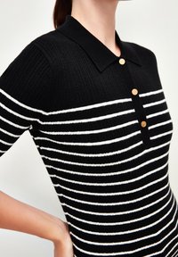 Polo rayé noir et blanc à côtes, avec manches courtes, col, et trois boutons dorés sur le devant.