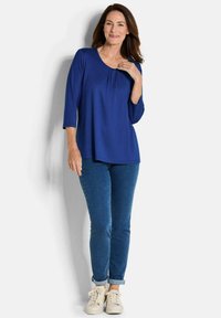 Dunkelblaues Oberteil mit drei Viertel Ärmeln, Rundhalsausschnitt und Falten-Details, kombiniert mit engen Jeans und weißen Sneakers.