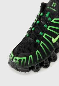 Black Nike Shox sneaker with green accents, featuring a mesh upper, distinctive curved design, and segmented sole for cushioning.