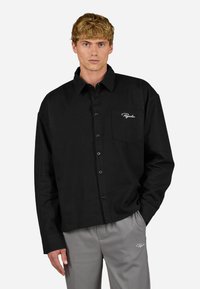 Camisa preta de botão, mangas longas, com um único bolso no peito com logo. Tecido suave, corte descontraído, botões visíveis, usada com calças cinza claro.