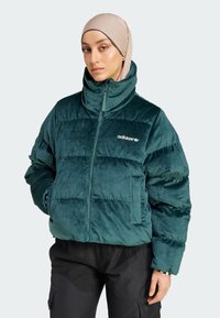 Kvinna i beige hijab och teal quiltad Adidas-jacka med hög krage, klädd i svarta byxor, stående med händerna i fickorna.