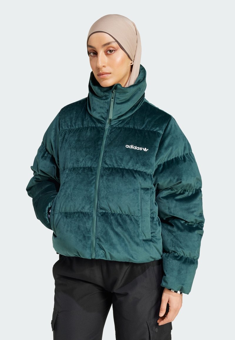 Frau mit beigem Hijab und teal-farbener, gesteppter Adidas-Jacke mit hohem Kragen und schwarzen Hosen, die mit den Händen in den Taschen steht.