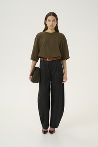 Mujer con blusa oversize verde oliva, pantalones anchos negros, cinturón marrón, sosteniendo un bolso de mano verde oliva y zapatos puntiagudos burdeos, de pie sobre fondo blanco.