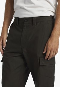 Dark green cargo pants made of durable fabric, featuring a button closure, side pockets, and a relaxed fit. Textured surface visible.