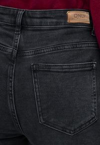 Джоб на черни дънки с бежова кожена етикета с надпис "ONLY Blue Denim" на червен плат фон.