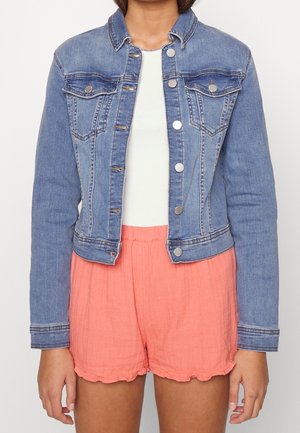 Femme portant une veste en jean bleu clair sur un haut blanc, assortie à un short en tissu froissé rose corail.