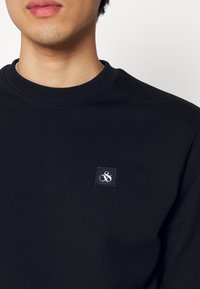 Suéter negro de cuello redondo hecho de una tela suave y texturizada. Presenta un pequeño parche cuadrado con un logo en azul marino y bordado blanco.