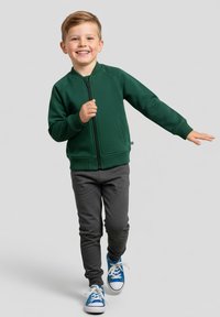 Zielona bluza na zamek z okrągłym kołnierzem, zestawiona z szarymi spodniami i niebieskimi tenisówkami. Tkanina wydaje się miękka, o gładkiej fakturze.