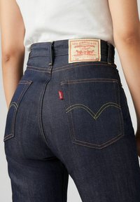 Ciemnoniebieskie dżinsy mają wysoką talię, żółte przeszycia oraz dwie tylne kieszenie z charakterystycznym kształtem V. Widoczna metka Levi's.