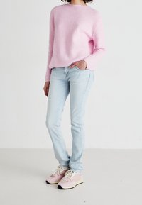 Osoba ubrana w jasnoróżowy sweter, jasnoniebieskie dżinsy i różowe trampki, stojąca z jedną ręką w kieszeni na białym tle.