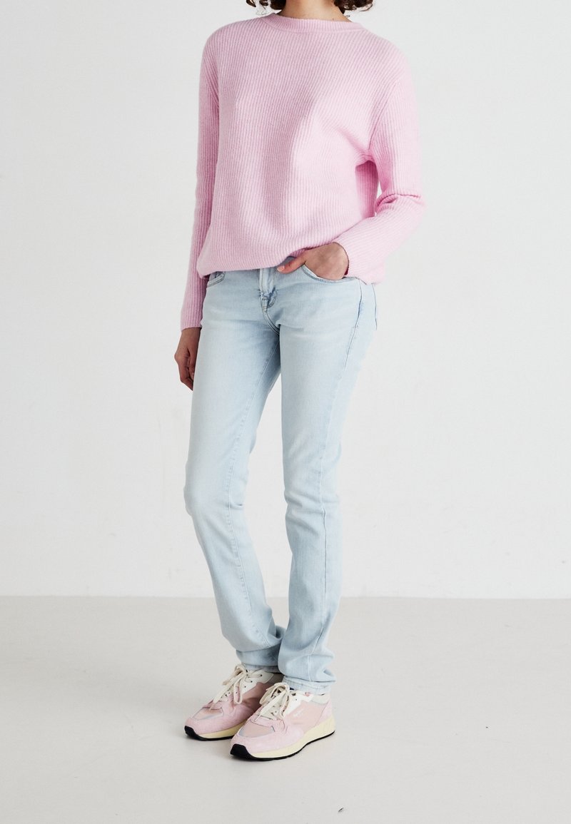 Osoba ubrana w jasnoróżowy sweter, jasnoniebieskie dżinsy i różowe trampki, stojąca z jedną ręką w kieszeni na białym tle.