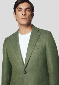 Zielony blazer o fakturze z pojedynczym zapięciem na guzik, z klapami i kieszenią na piersi, noszony na białej koszulce.