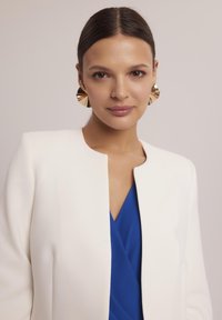 Biała marynarka z okrągłym kołnierzem, zestawiona z niebieską bluzką z dekoltem w kształcie litery V. Duże, złote geometryczne kolczyki podkreślają całość. Gładkie faktury we wszystkich elementach.