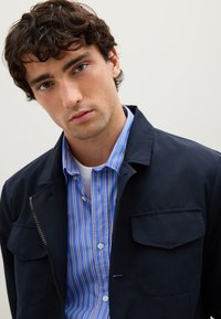 Junger Mann mit lockigem braunem Haar, der eine marinefarbene Jacke über einem blau-weißen gestreiften Hemd trägt und direkt in die Kamera schaut.