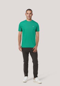 Grünes Baumwoll-T-Shirt mit Rundhalsausschnitt und kurzen Ärmeln, kombiniert mit dunklen Hosen und weißen Sneakers. Keine sichtbaren Muster oder Akzente.