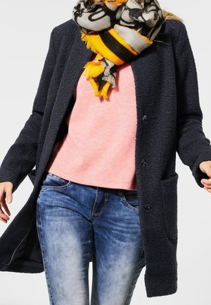 Marineblauwe textuurjas met zakken, gedragen over een lichtroze trui, gecombineerd met blauwe denimjeans en een gemêleerde sjaal in grijs, zwart en geel.