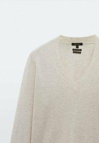 Beige V-neck sweater made of soft knit fabric, featuring ribbed cuffs and a smooth texture. Label visible inside the neck opening.