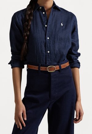 Woman wearing a dark blue button-up shirt with rolled sleeves, dark blue pants, and a brown leather belt with gold buckle.