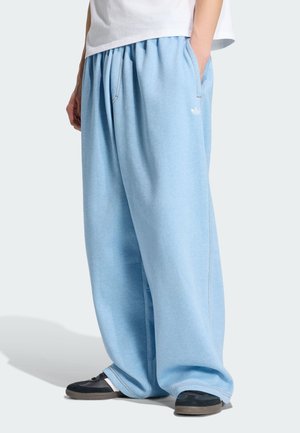 Persona che indossa pantaloni della tuta azzurri chiari e morbidi con tasche laterali e scarpe da ginnastica nere, in piedi con le mani nelle tasche.