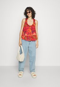 Top de tanque vermelho com padrão, decote redondo, combinado com jeans azul claro e sandálias de slide brancas e fofas. Segura uma bolsa de mão suave em creme.