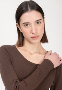 Brauner langärmeliger Pullover mit weitem Halsausschnitt, gerippten Bündchen und glatter Textur. Eine zarte Halskette mit einem kleinen Anhänger ist sichtbar.