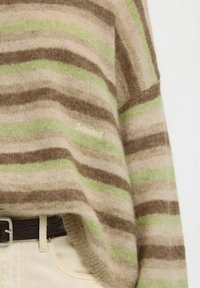 Pull rayé doux en tons beige, marron et vert clair, porté sur un pantalon crème avec une ceinture marron foncé, présentant une légère texture et un texte de marque.