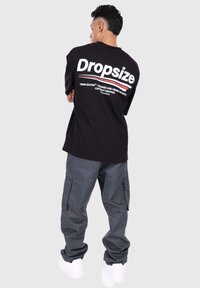 Black t-shirt with white "Dropsize" text on the back, paired with gray cargo pants and white sneakers, viewed from the back.