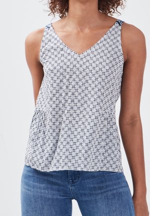 Woman wearing sleeveless white top with small black dot pattern and blue jeans, against a plain light background.