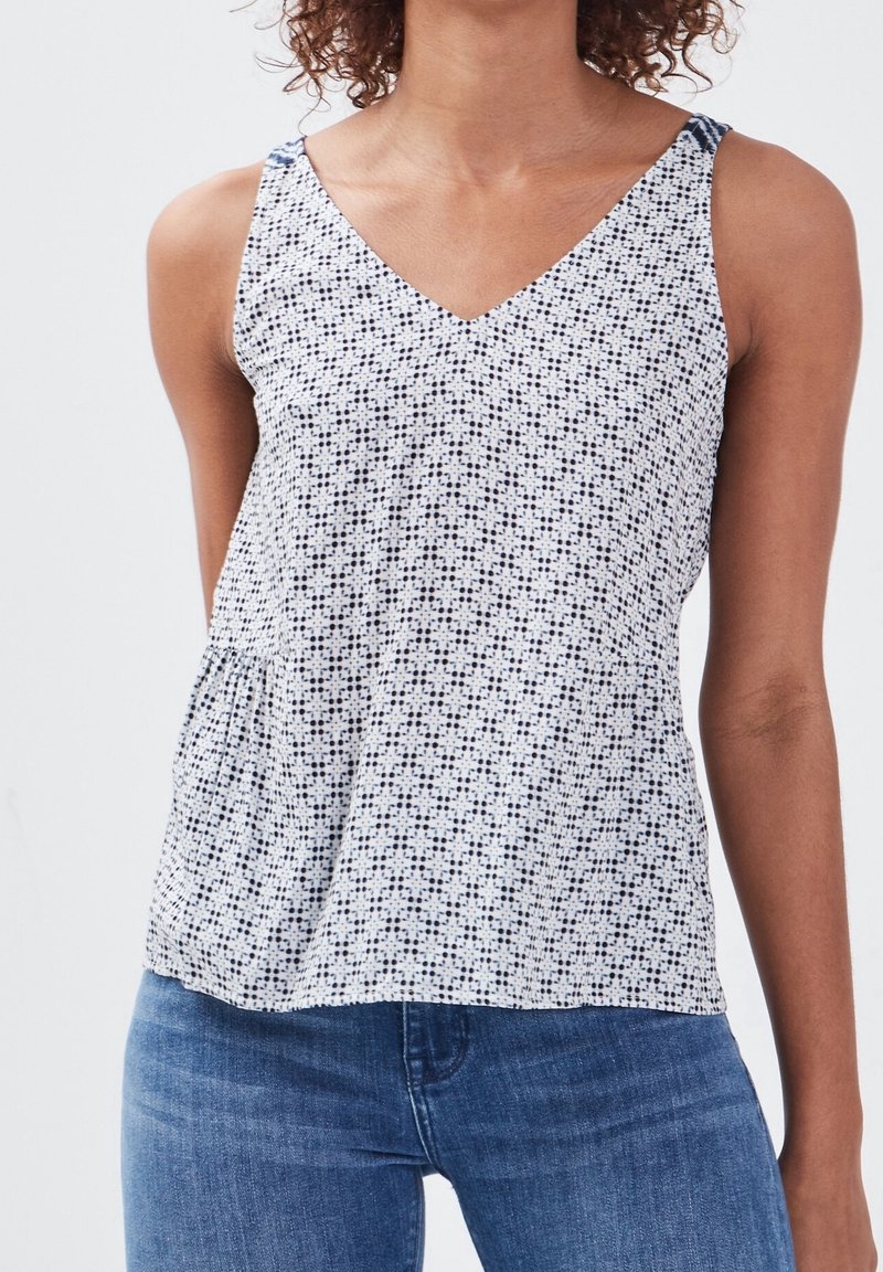 Femme portant un haut blanc sans manches avec un petit motif à pois noirs et un jean bleu, sur un fond clair uni.