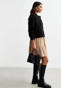 Black bomber jacket with snap buttons, beige tiered dress, tall black leather boots, and a small textured black handbag.