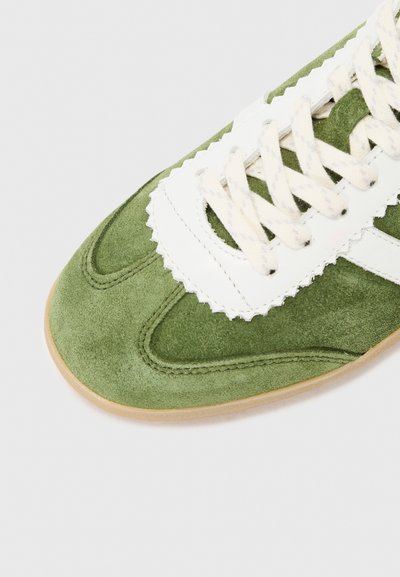 Roheline sametist sneaker valgete nahkakreemide, lainelise disaini ja kreemjatest seotud paeladest. Omab kaardus õmblusi ja heledat kummitalda.