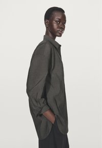 Dunkelgraues Hemd mit Kragen und langen Ärmeln, lockerer Passform, mit einer dezenten Textur und abgerundetem Saum, getragen von einem Model.