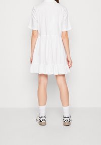 White cotton short-sleeve dress with a ruffled hem, collar detail, and a relaxed fit, paired with sporty black and white sneakers.