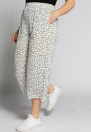 Femme portant un pantalon court blanc à pois noirs, une main dans la poche, associé à des baskets blanches et un haut noir.