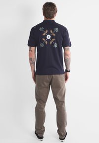 Homme avec des tatouages portant une chemise marine avec un design de croix florale dans le dos, un pantalon marron et des baskets noires, se tenant de dos.