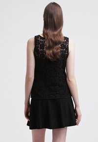 Top de encaje negro sin mangas con patrón floral y acentos redondeados, combinado con una falda negra plisada, visto desde atrás. Textura suave.